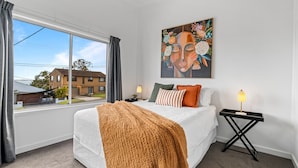 A bedroom with a large bed, a window with curtains, a nightstand with a lamp, and a painting on the wall. - Lakeside 2-bed Retreat With Sunny Balcony (Buff Point)