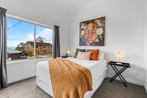 Quarto com uma cama grande, uma janela com cortinas, uma mesa de cabeceira com abajur e um quadro na parede.