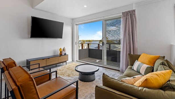 A modern living room with a flat-screen TV, leather furniture, a round coffee table, and a sliding glass door leading to an outdoor balcony. - Lakeside 2-bed Retreat With Sunny Balcony (Buff Point)