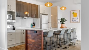 A modern kitchen with a central island, bar stools, wooden cabinets, and pendant lighting. - Lakeside 2-bed Retreat With Sunny Balcony (Buff Point)