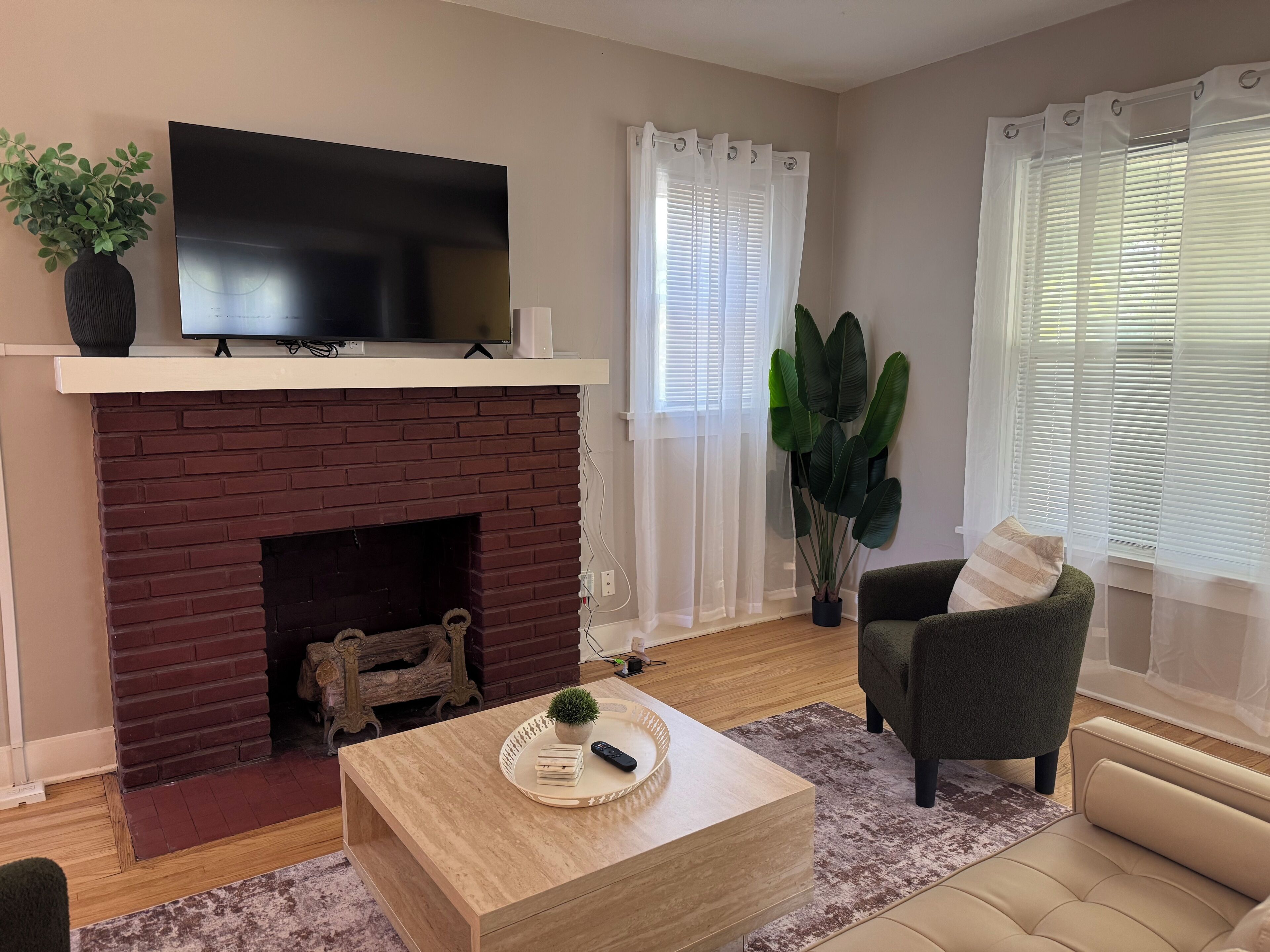 A living room with a brick fireplace, a flat-screen TV mounted above it, a comfortable sofa, a coffee table with a tray, and a large plant.