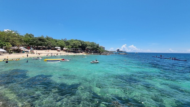 Une plage avec une eau turquoise et limpide, des bateaux et une rive sablonneuse, avec des bâtiments en arrière-plan.