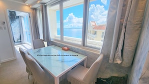 Un espace repas avec une table et des chaises en verre, de grandes fenêtres offrant une vue sur l’océan et une vue sur les bâtiments situés de l’autre côté de l’eau.