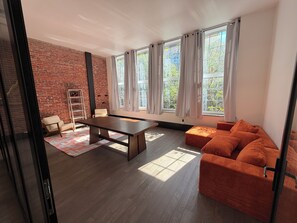 Ein modernes Wohnzimmer mit Backsteinwand, großen Fenstern mit Vorhängen, einem Holztisch, einer Couch und einem Sessel.
