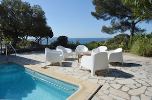 Ein Poolbereich mit weißen Stühlen und einem Tisch, freier Blick auf das Meer und eine terrasse mit Steinplatten.