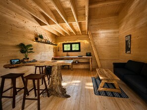L’intérieur d’un chalet en bois comprend une table à manger, des tabourets de bar, un canapé noir, un coin cuisine et une fenêtre donnant sur un paysage agréable.