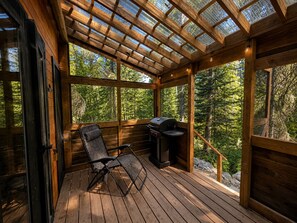 Terraza de madera con una parrilla y una silla, rodeada de paneles de vidrio y vistas al bosque.