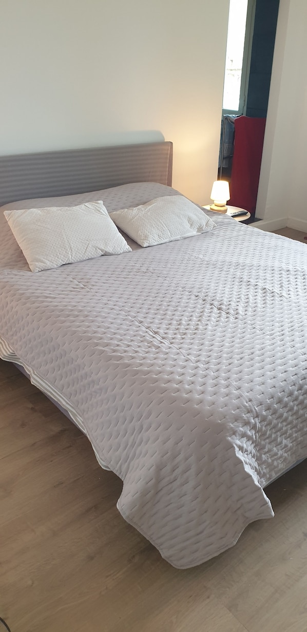 A neatly made bed with a textured white duvet, a wooden headboard, and a bedside table with a lamp. - Typical Corsican village. (Sant’Antonino)
