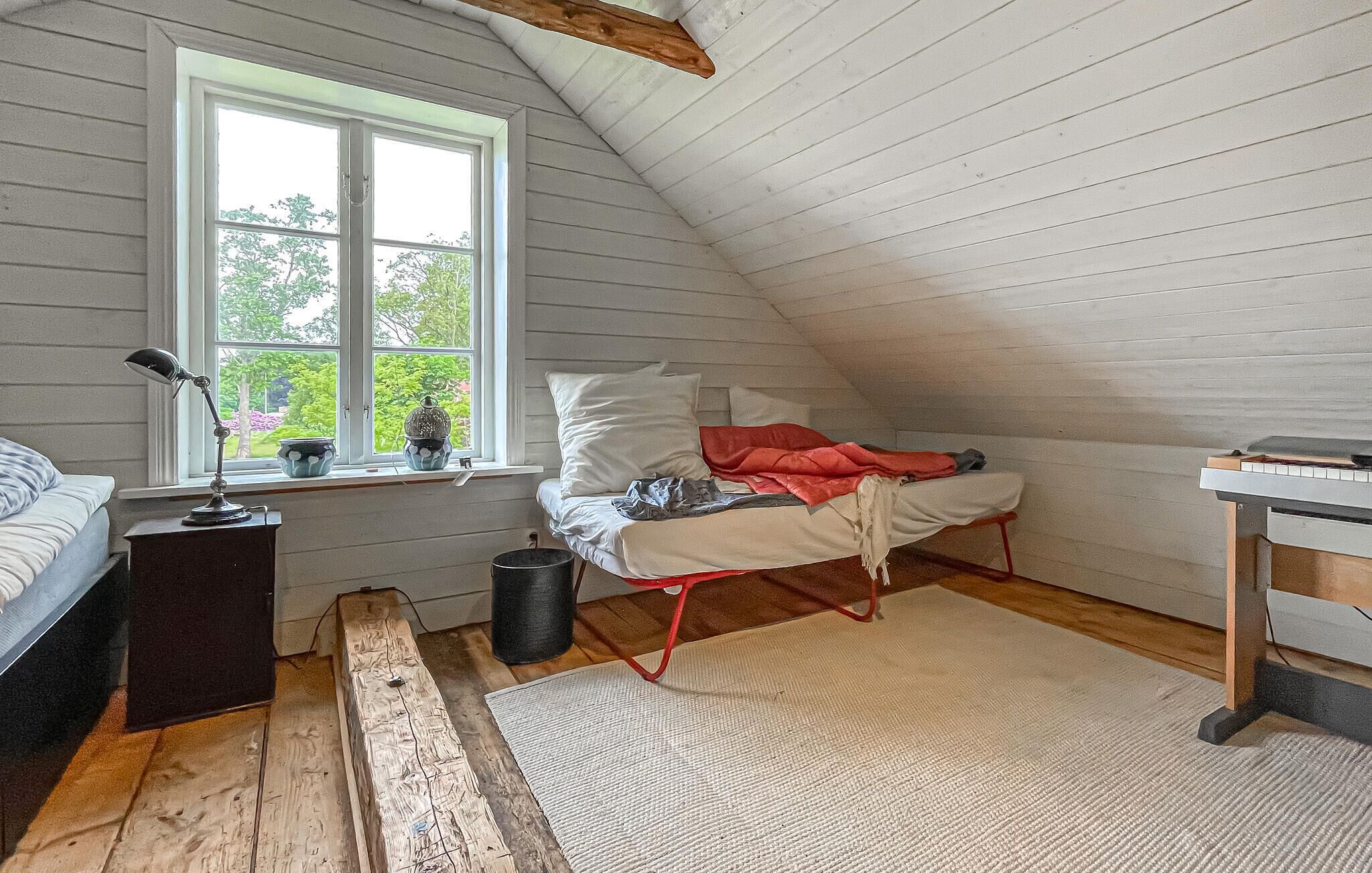 Une pièce avec un plafond en pente, un lit avec des oreillers et une couverture, un bureau avec une chaise, une fenêtre avec des plantes et une lampe sur une table de chevet.