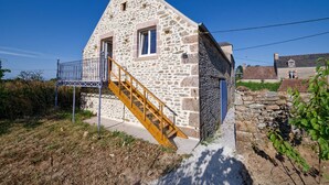 A stone house with a wooden staircase leading to a door, surrounded by vegetation and another building in the background. - Balcony Cottage (Varouville)