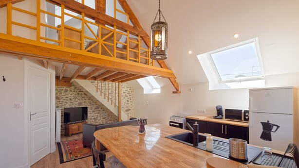 A kitchen with a wooden table, a stone wall, a TV, and a view of the sky. - Balcony Cottage (Varouville)