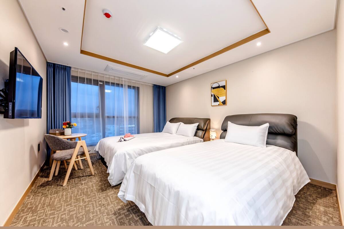 A hotel room with two beds, a desk, a chair, a TV, and a window with curtains.