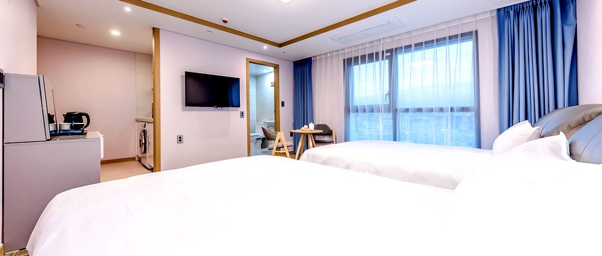 Habitación de hotel con dos camas, televisión, una mesita y vista a la ciudad desde la ventana.
