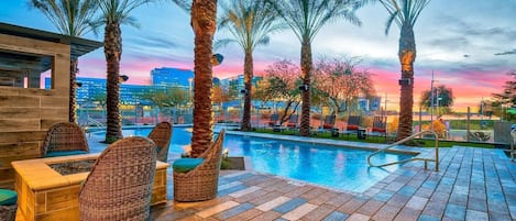 A poolside area with wicker furniture and palm trees, overlooking a cityscape at sunset.