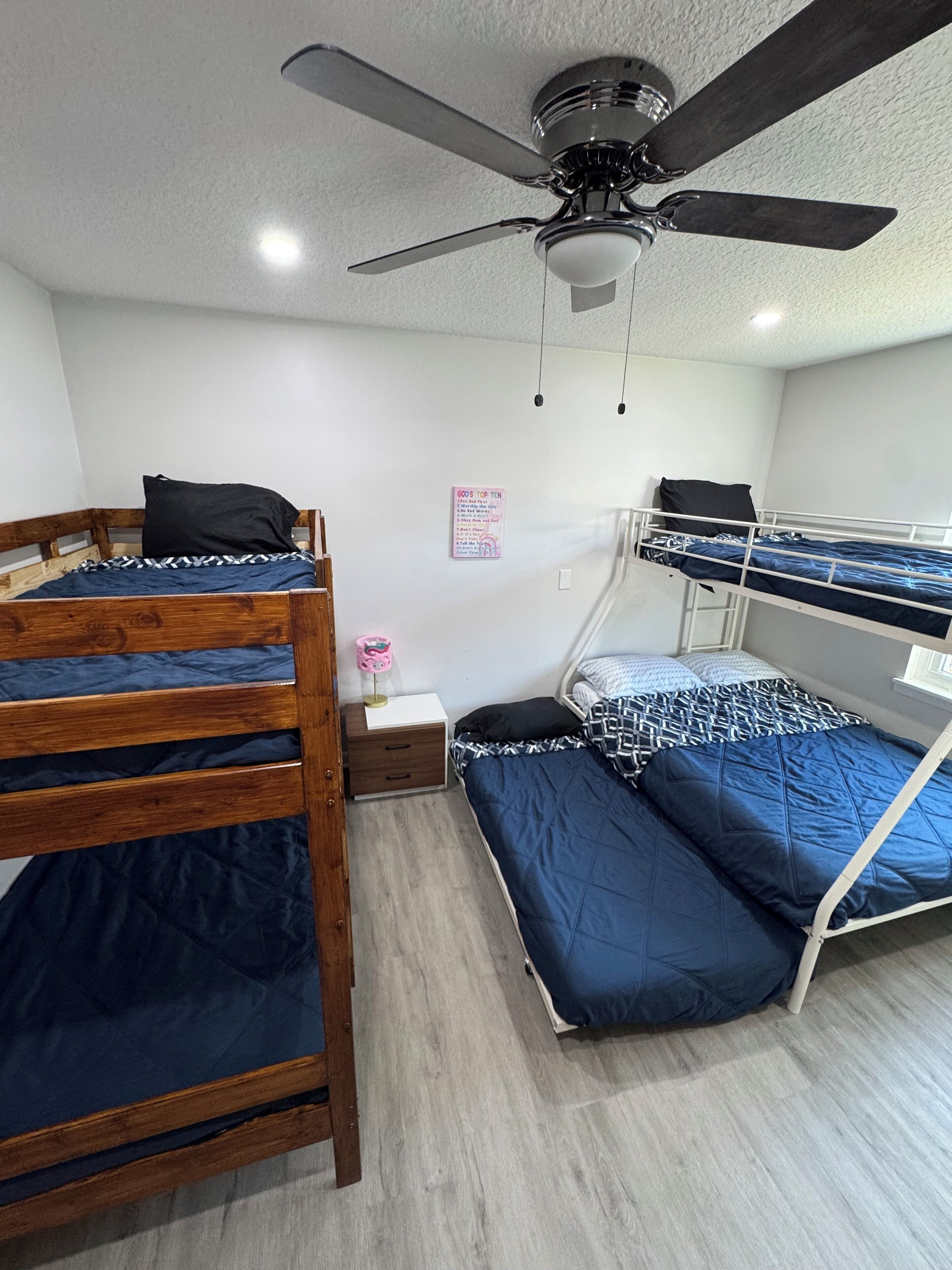 Habitación con literas, un ventilador de techo, una mesita de noche con lámpara y un cartel en la pared.