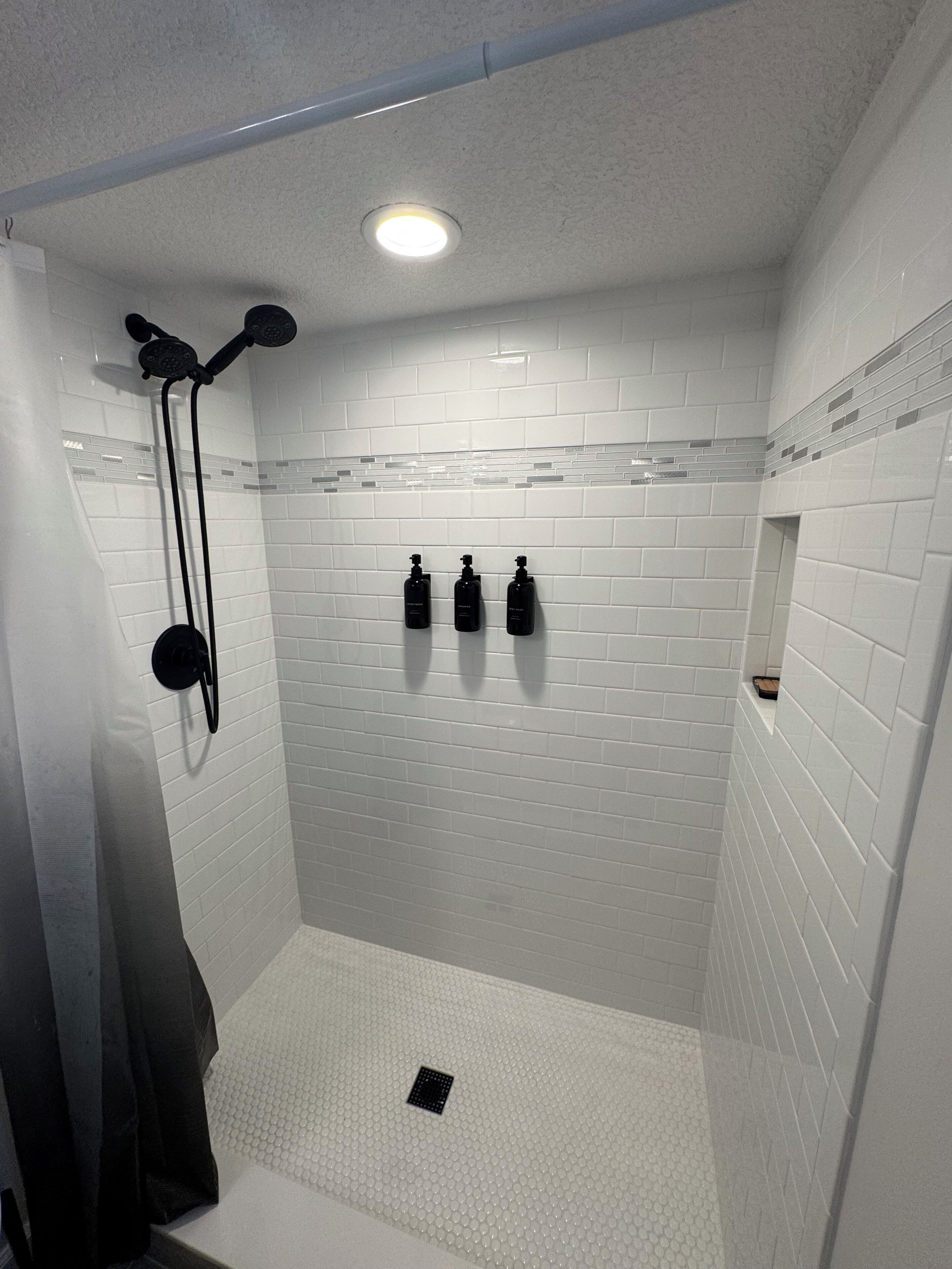Eine Dusche mit weiß gefliesten Wänden, einer Deckenleuchte und drei schwarzen Flaschen an der Wand.