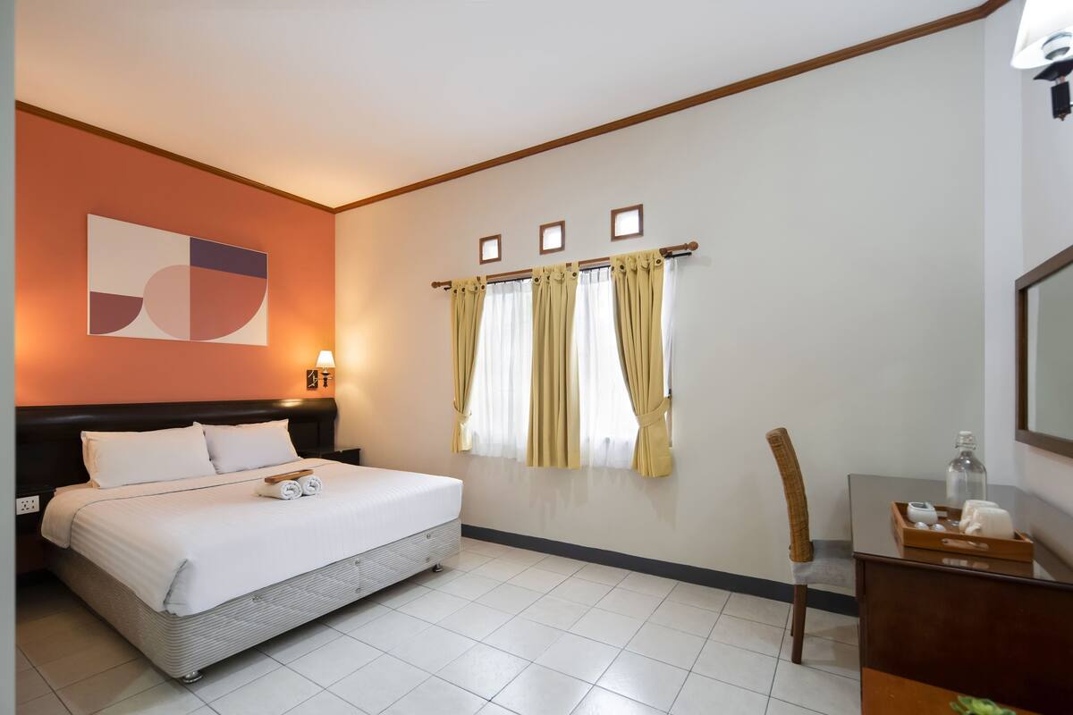 Ein Hotelzimmer mit Bett, Schreibtisch, Stuhl, einem Fenster mit Vorhängen und einem Wandbild.