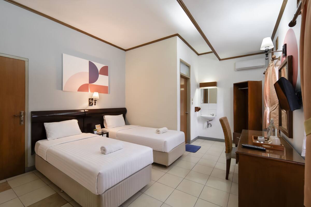 Ein Hotelzimmer mit zwei Betten, einem Schreibtisch, einem Stuhl, einer Lampe, einem Spiegel und einem durchgegangenen Tür erkennbaren Badezimmer.