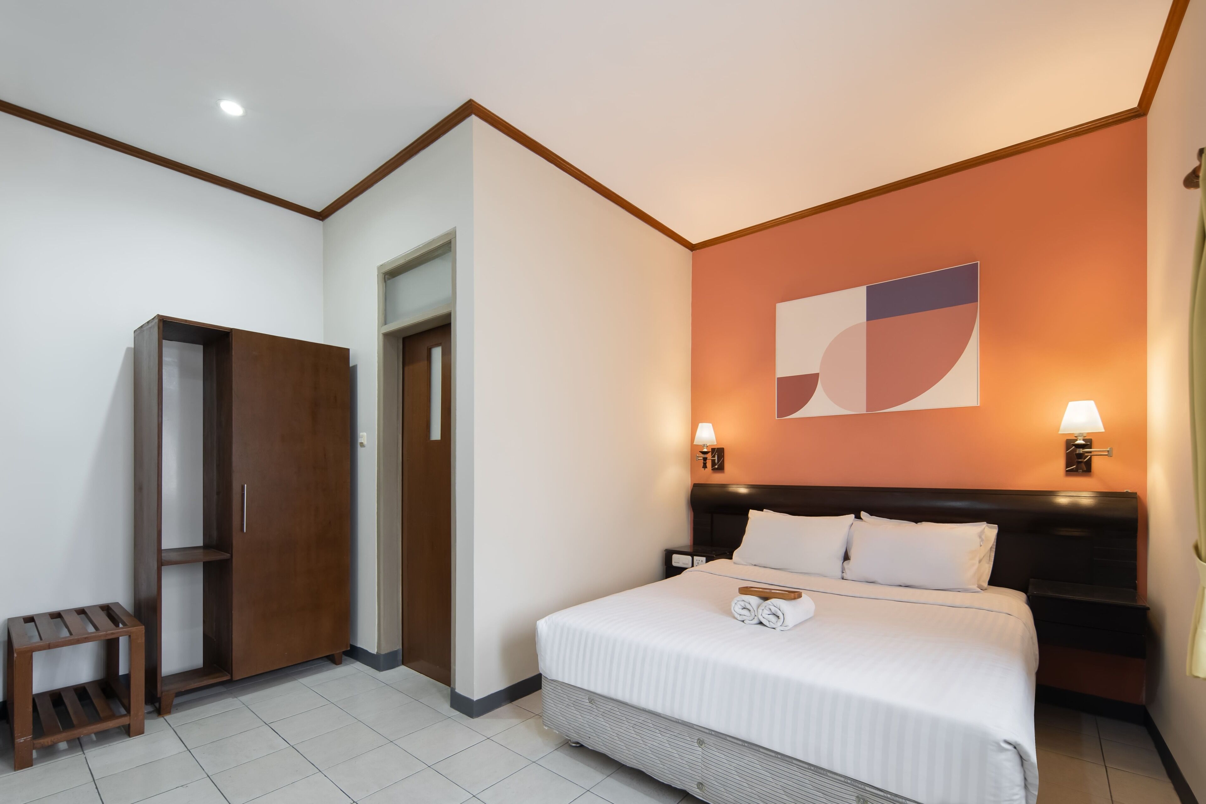 Quarto de hotel com cama, mesas de cabeceira, guarda-roupa, um pequeno banco e uma parede com uma obra de arte geométrica.