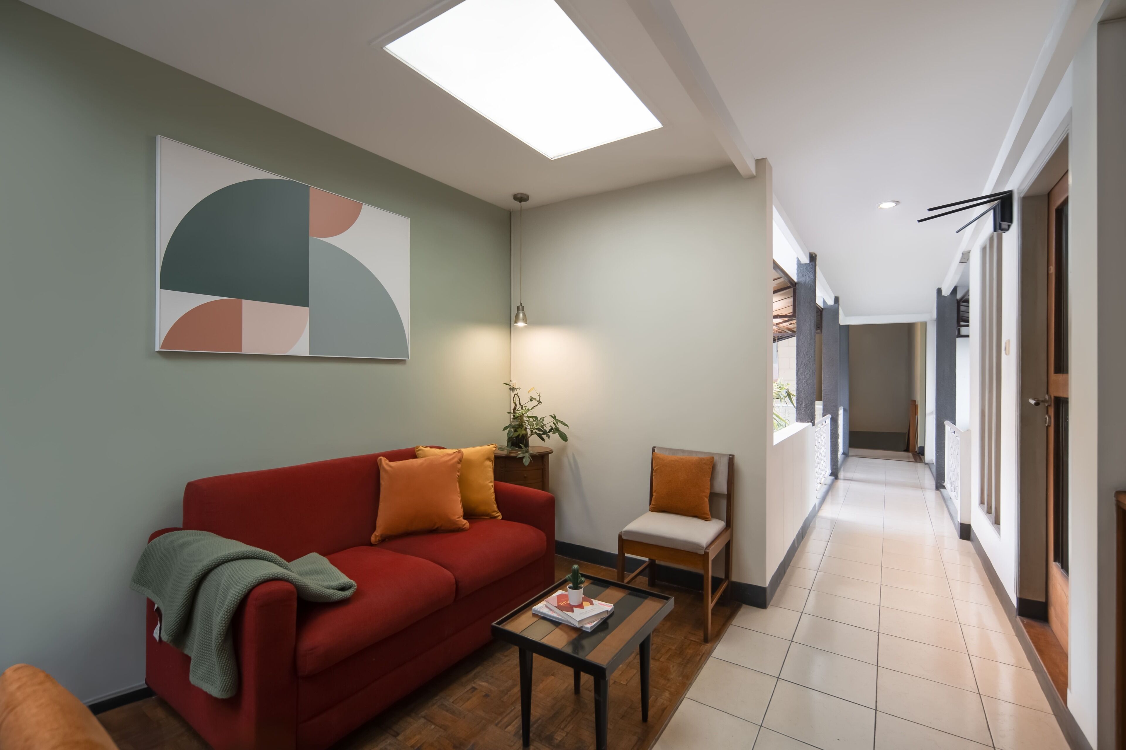 A modern living room with a red sofa, a small table, and a chair. There is a skylight above the sofa and a hallway leading to other rooms.