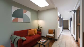 A modern living room with a red sofa, a small table, and a chair. There is a skylight above the sofa and a hallway leading to other rooms.
