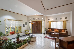A spacious living room with a sofa, a coffee table, and a dining table. There are potted plants and a window with curtains.