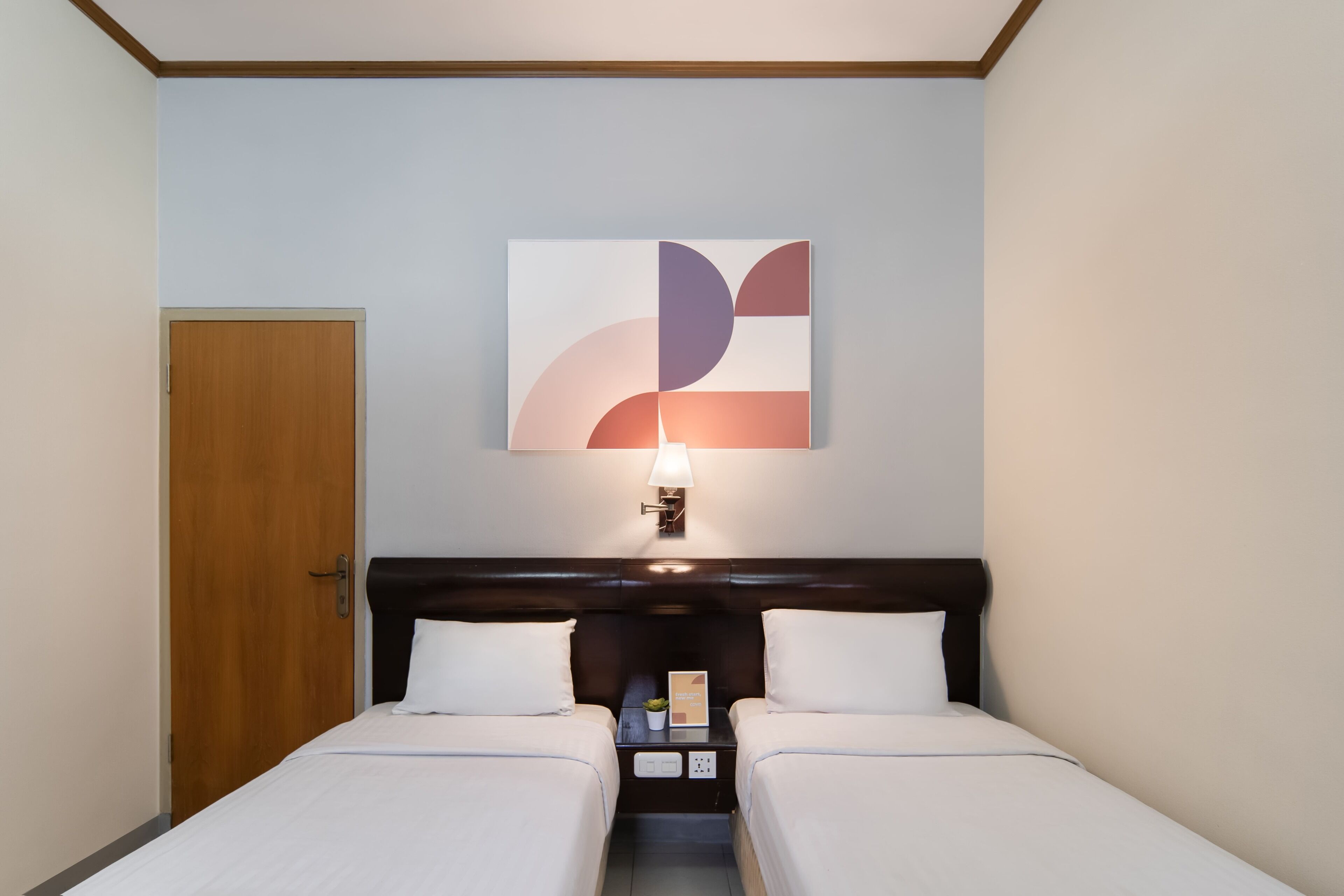 Habitación de hotel con dos camas, una puerta de madera, una obra de arte colgada en la pared y una mesita de noche con lámpara.