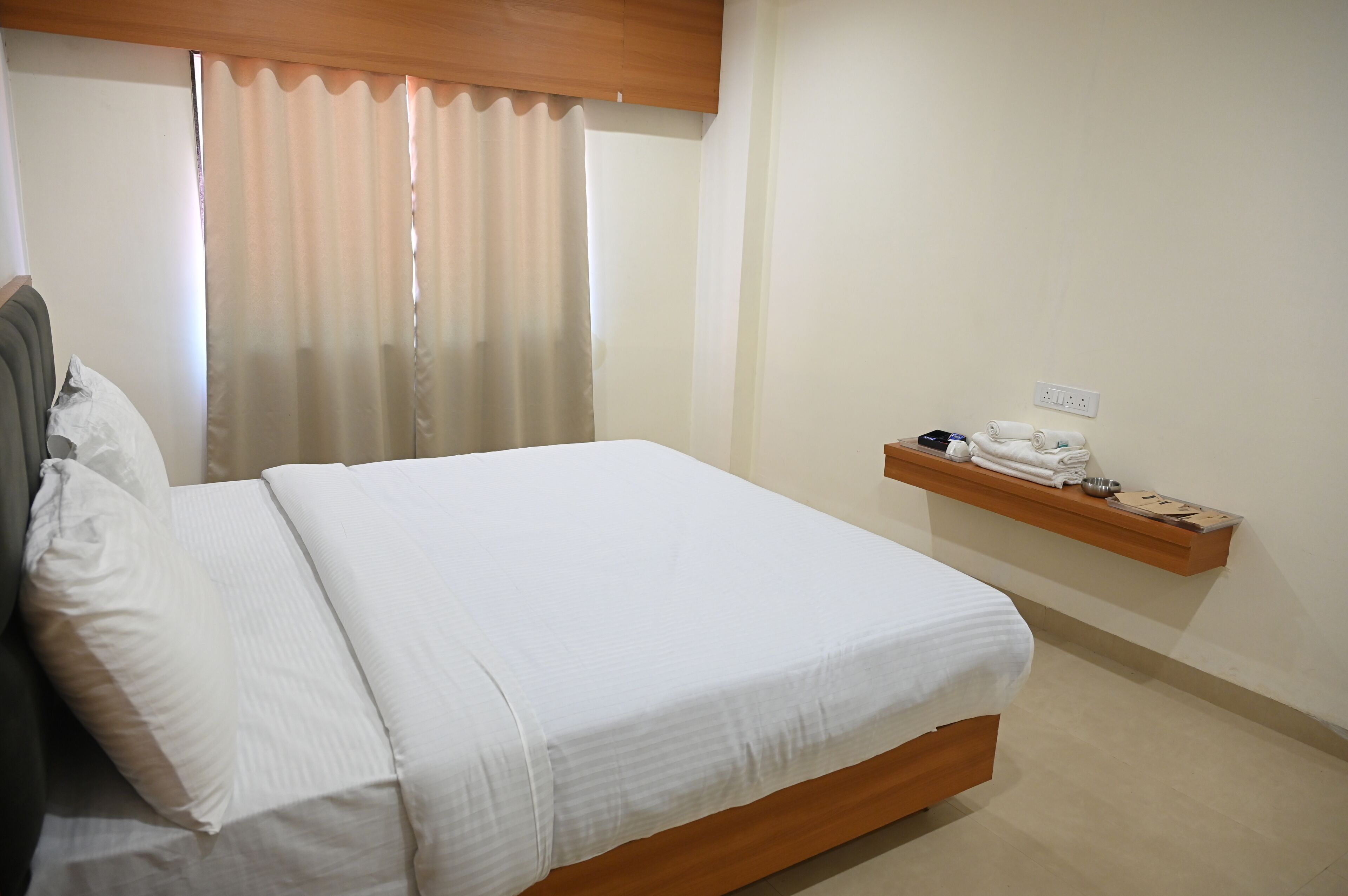 Una cama bien hecha con cabecera de madera, una mesita de noche con toallas y artículos de aseo, y una ventana con cortinas.