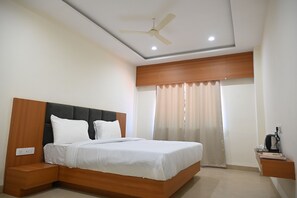 A hotel room with a bed, a nightstand, a ceiling fan, and a window with curtains. - Shubhratna Hotels & Resort (Barnagar)