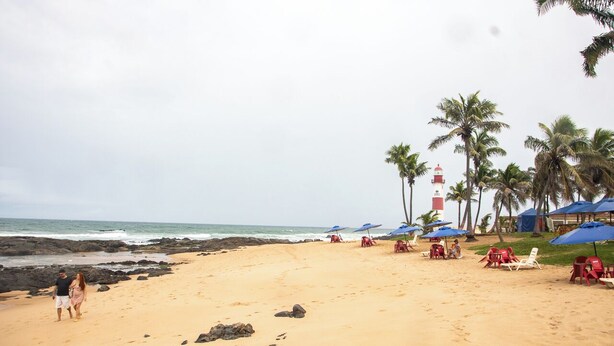 A beach with chairs and umbrellas, a lighthouse, and palm trees. - Ed. Studio Oceânico (Salvador)