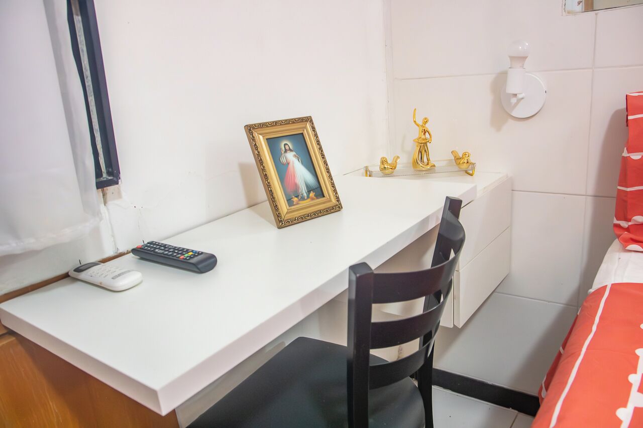 Ein Zimmer mit einem weißen Schreibtisch, einem schwarzen Stuhl, einem gerahmten Bild, einer Fernbedienung und einer kleinen Statue auf einem Regal.