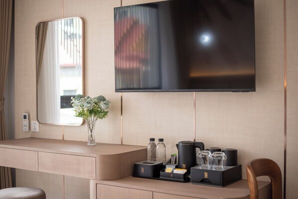 A hotel room with a flat-screen TV mounted on the wall, a mirror, a wooden desk with a vase of flowers, and a coffee maker. - Thadatra Inn (Phnom Penh)
