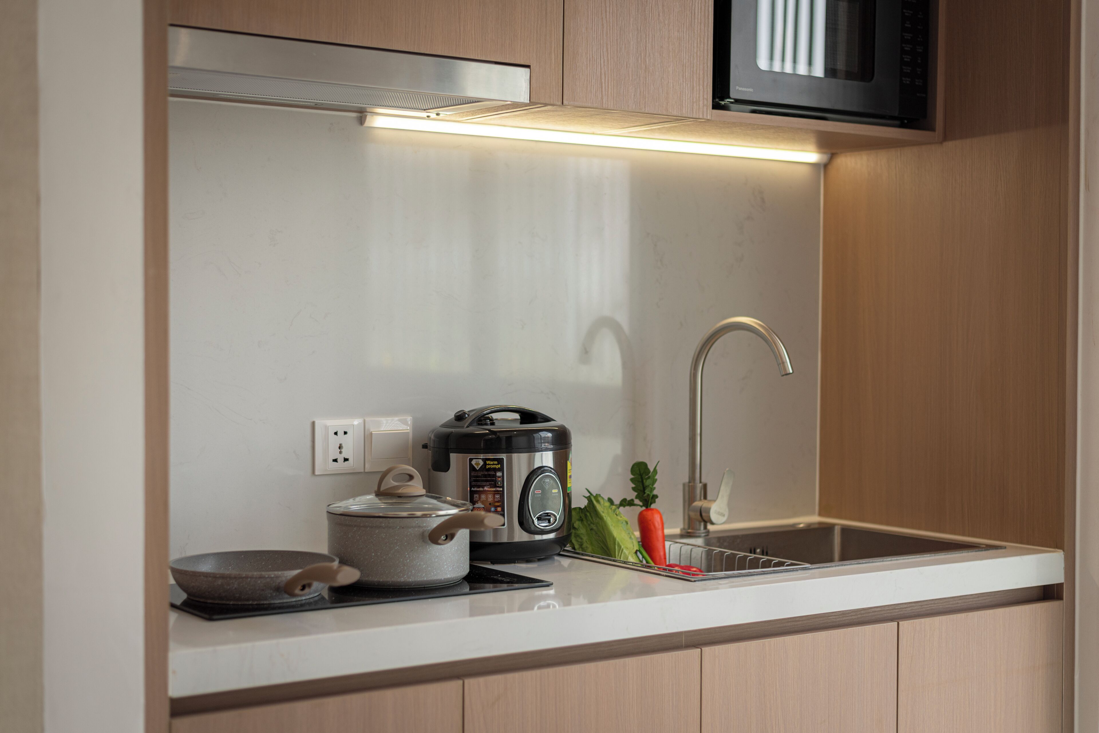Una cocina con un microondas sobre una campana extractora de acero inoxidable, una cocedora de arroz, una sartén y un lavabo con grifo.