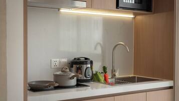 Una cocina con un microondas sobre una campana extractora de acero inoxidable, una cocedora de arroz, una sartén y un lavabo con grifo.