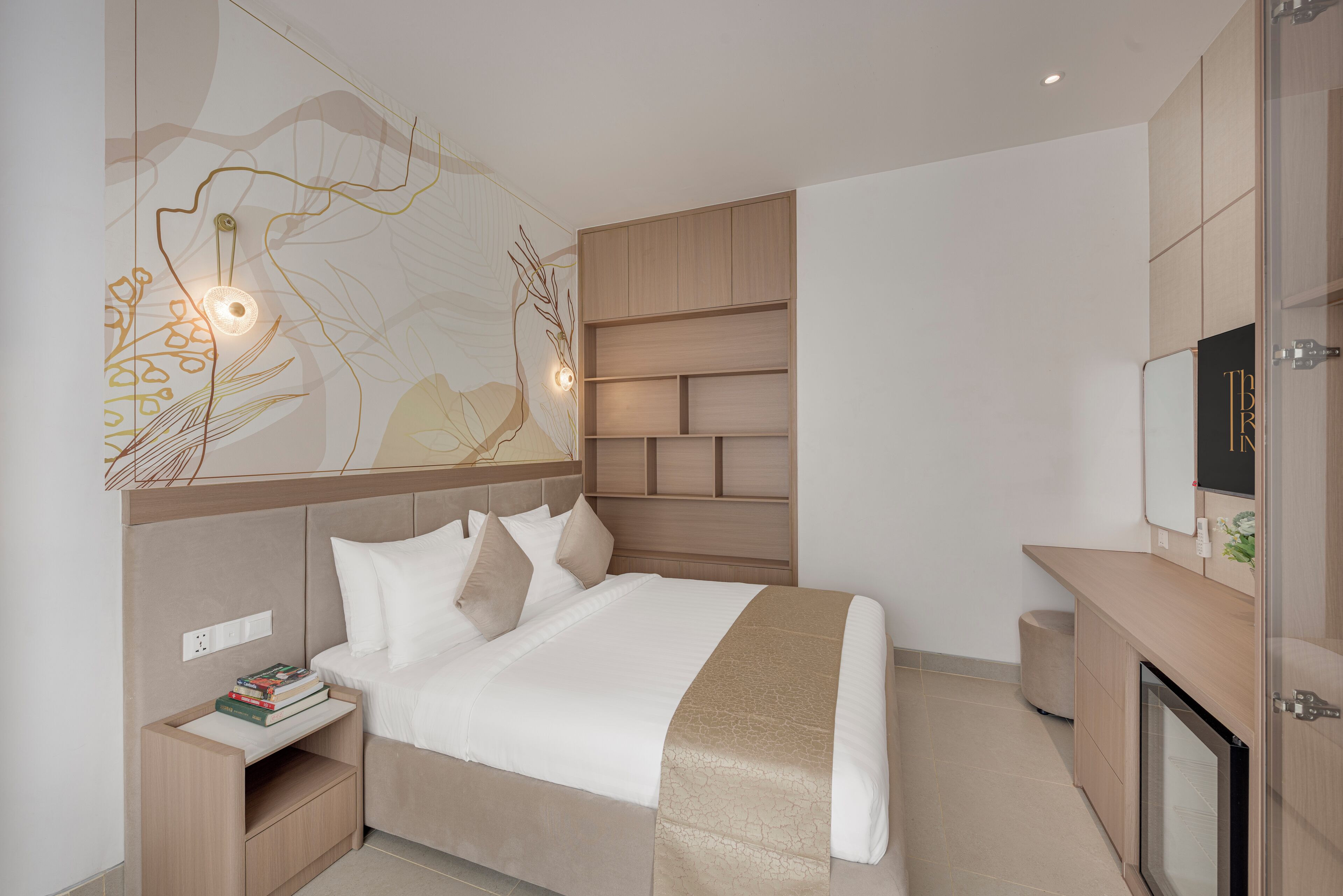 A modern hotel room with a bed, bedside tables, a nightstand, a wall-mounted TV, and a built-in shelving unit.