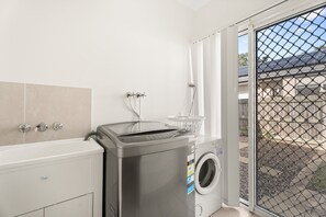 A laundry area with a washing machine, dryer, and a window with a view of a backyard. - Coastal 4-bed Retreat With Pool in Toogoom (Toogoom)