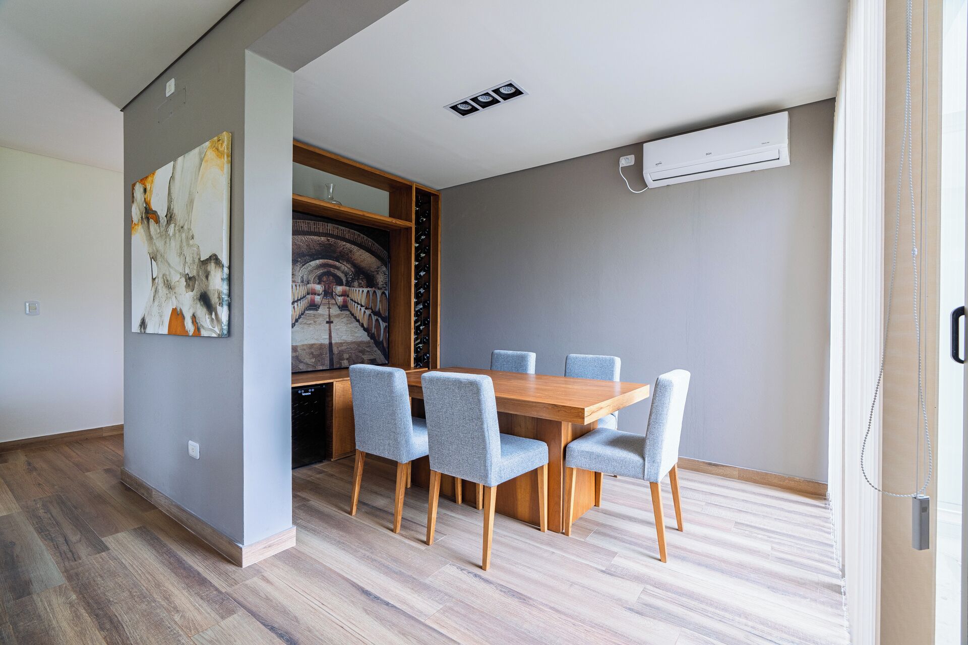 A dining area with a wooden table and four chairs, a wine rack, and a painting on the wall.