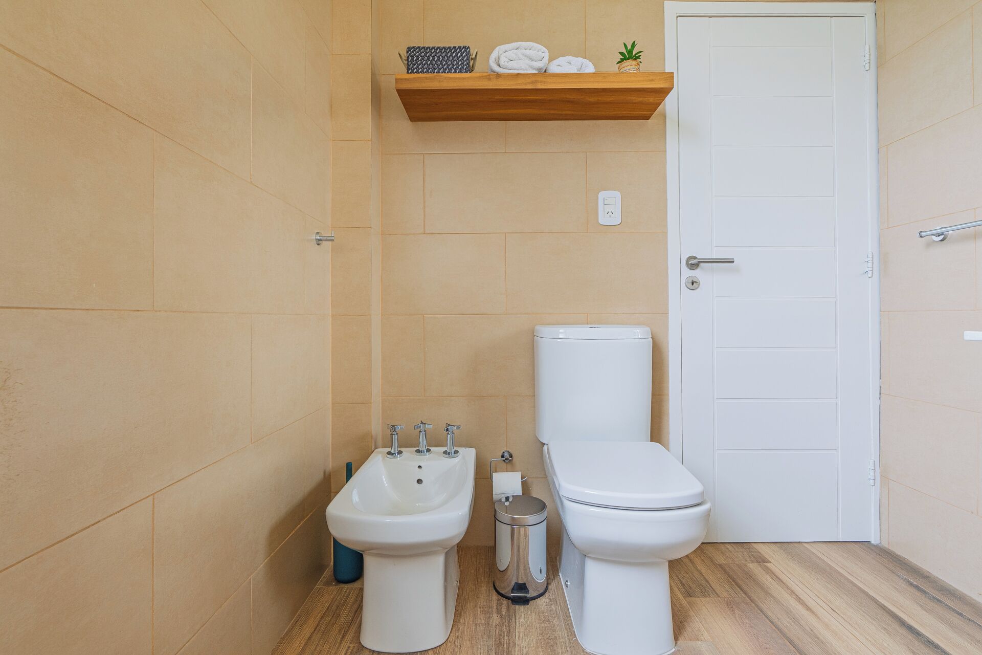 Un baño con paredes de azulejos color beige, un inodoro blanco, un bidé y un estante con toallas y una planta.