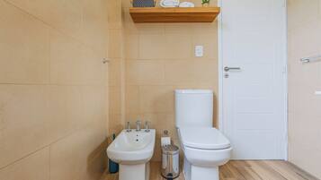 Un baño con paredes de azulejos color beige, un inodoro blanco, un bidé y un estante con toallas y una planta.