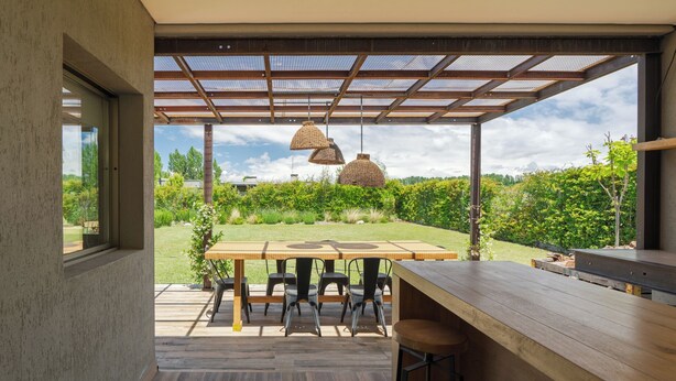 Un área de comedor al aire libre de diseño moderno, con mesa y sillas de madera, una pérgola con techo de malla y lámparas colgantes.