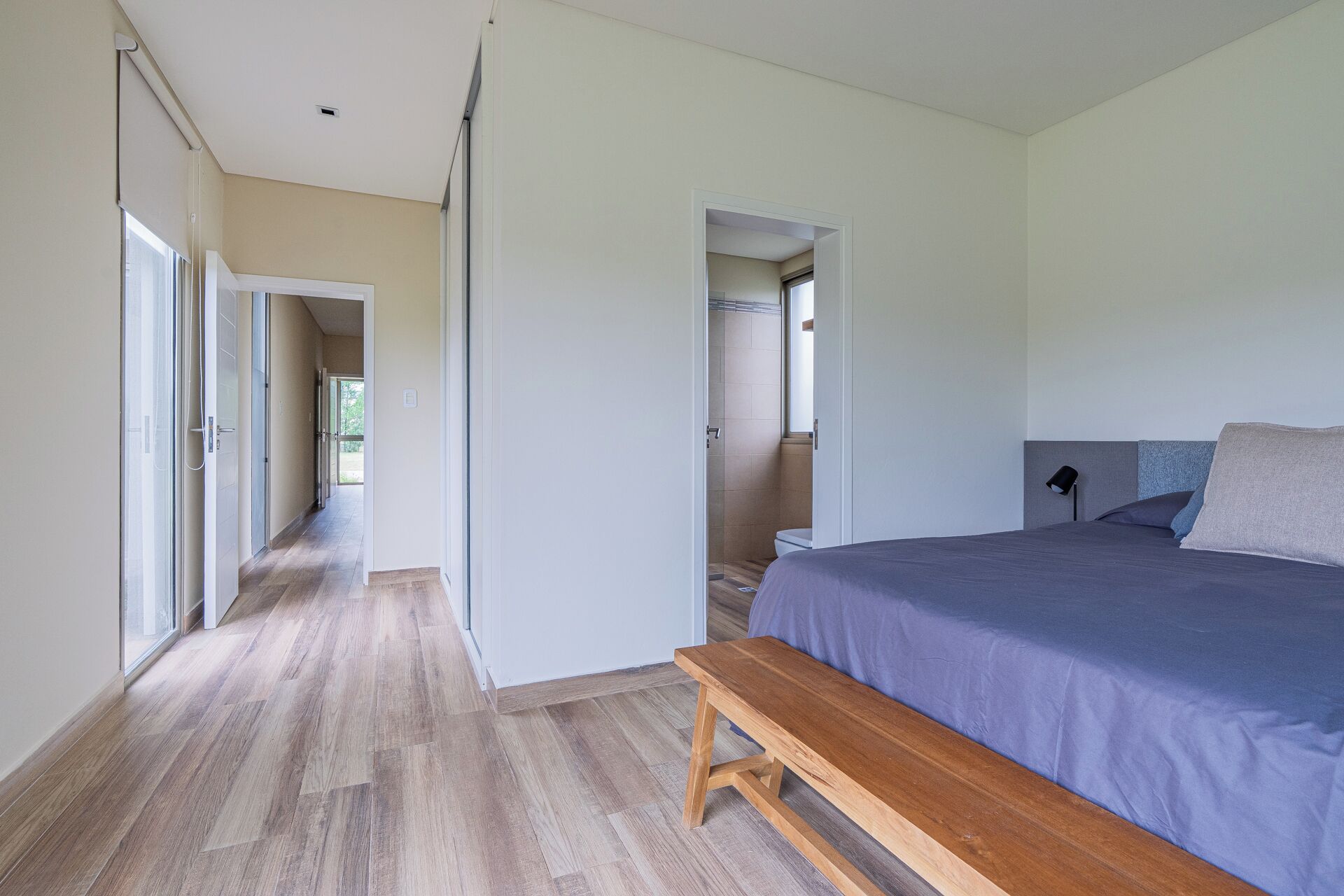 A bedroom with a bed, a bench, a bathroom with a shower, and a hallway leading to an outdoor area.