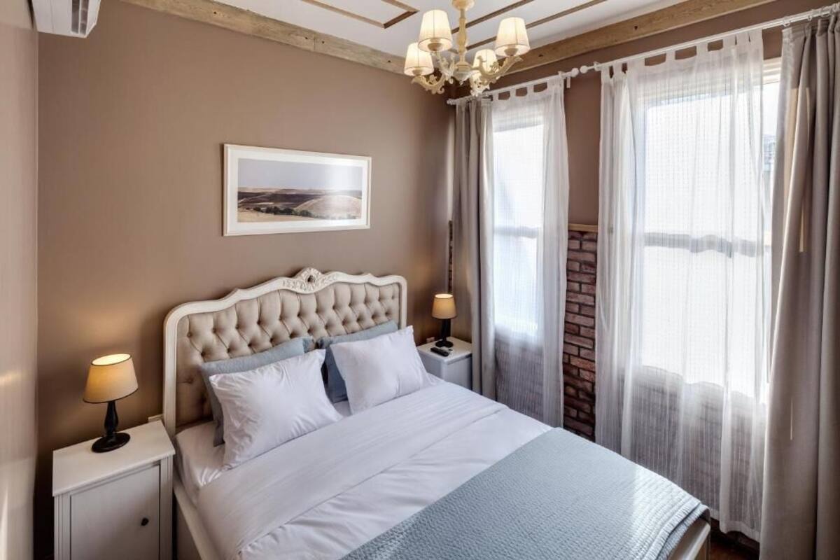 Una camera da letto con una testiera trapuntata, un letto con lenzuola bianche, un comodino con una lampada, un quadro appeso alla parete e una finestra con tende leggere.