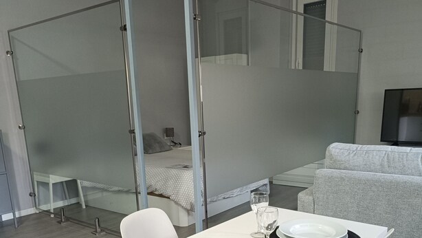 A modern, minimalist room with a white table set for two, a grey sofa, and a bed with a glass partition. - Palacio Congresos - Airport (Sevilla)