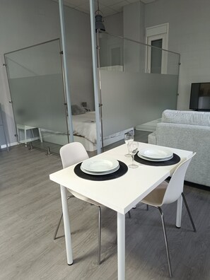Une chambre moderne et minimaliste, avec une table blanche dressée pour deux personnes, un canapé gris et un lit séparé par une cloison en verre.