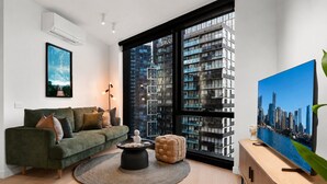 A modern living room with a green sofa, a round coffee table, a large window with a city view, and a flat-screen TV on a wooden console. - Skyline 2-bed Near Crown With Pool and Sauna (Melbourne)