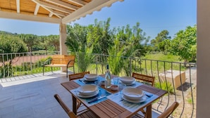 Un espacio de comedor al aire libre cubierto, con una mesa dispuesta para cuatro personas, rodeada de sillas y vistas a un paisaje exuberante.