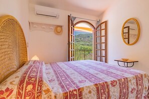 A bedroom with a bed, an air conditioning unit, a window with an open door, a balcony, and a small table with a bowl. - Villa Valentina - Villa Valentina 1 (Capoliveri)