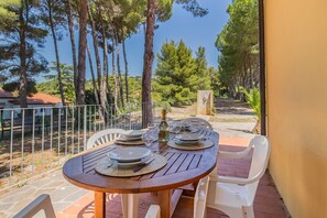 A wooden table with plates and glasses, surrounded by chairs, set on a patio with a view of trees and a stone wall. - Villa Valentina - Villa Valentina 2 (Capoliveri)