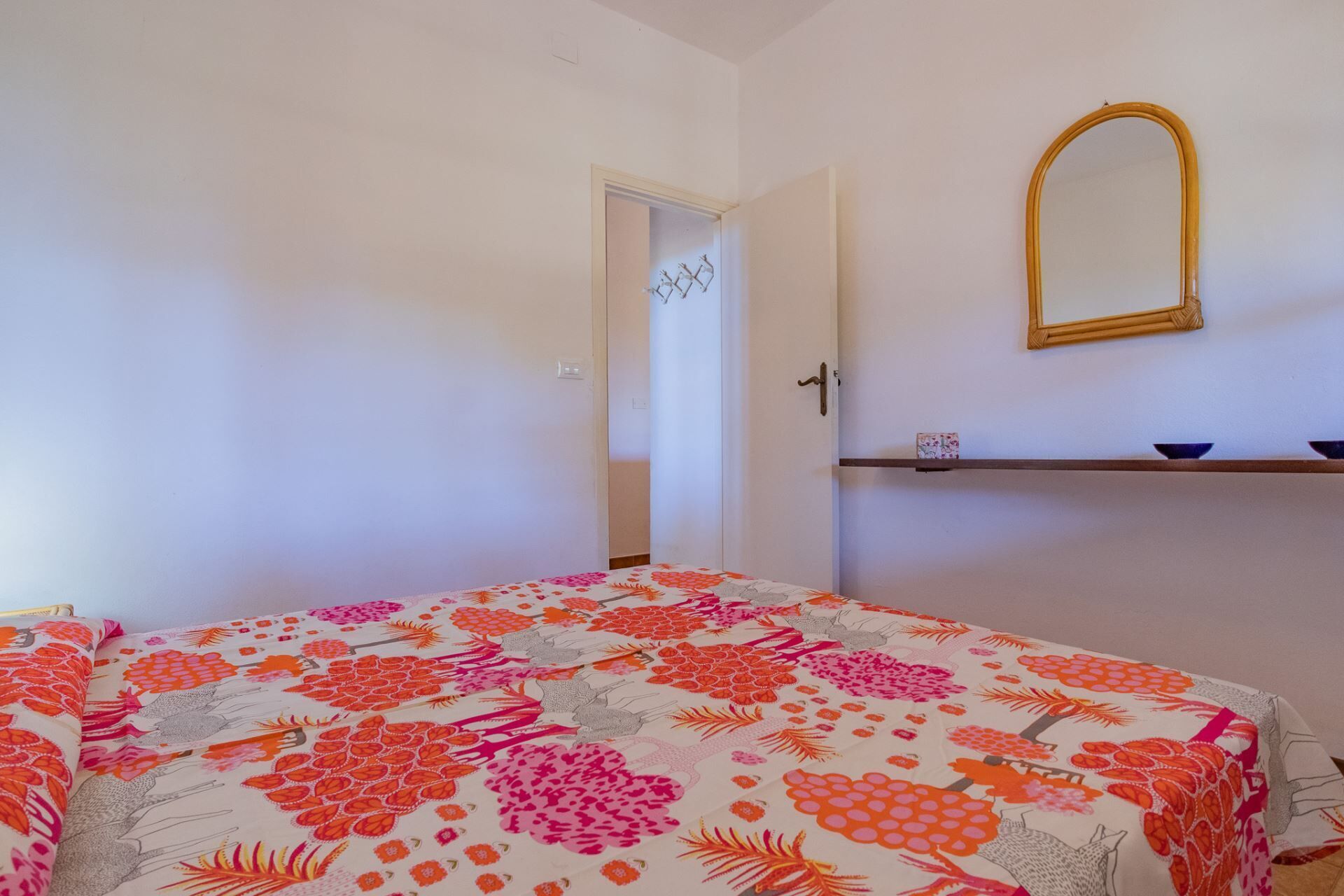 A bedroom with a bed, a door, a mirror, and a shelf.