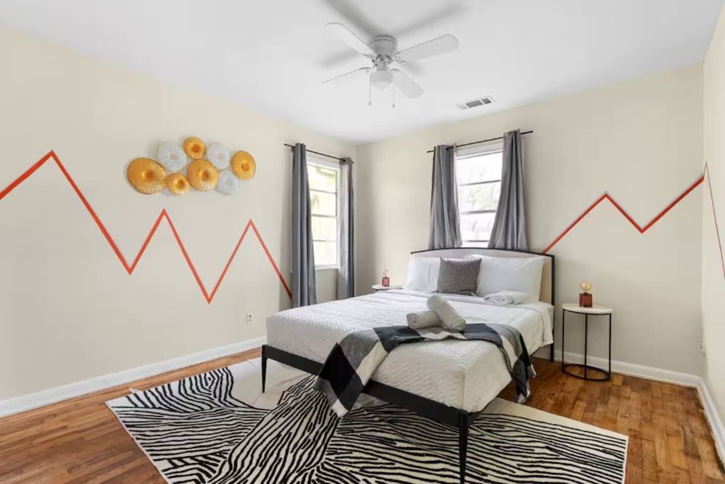 Une chambre à coucher comprenant un lit, un ventilateur de plafond, un tapis à motifs zébrés, une petite table ronde avec un globe terrestre et un mur orné d’un motif en zigzag.